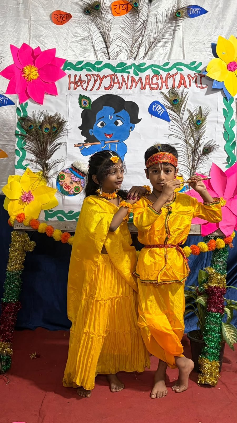 Students dressed as Radha and Krishna in front of the Happy Janmashtami board